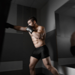 Male boxer training with punching bag in dark sports hall. Young boxer training on punching bag. Male boxer as exercise for the big fight. Boxer hits punching bag. Young masculine male athlete.